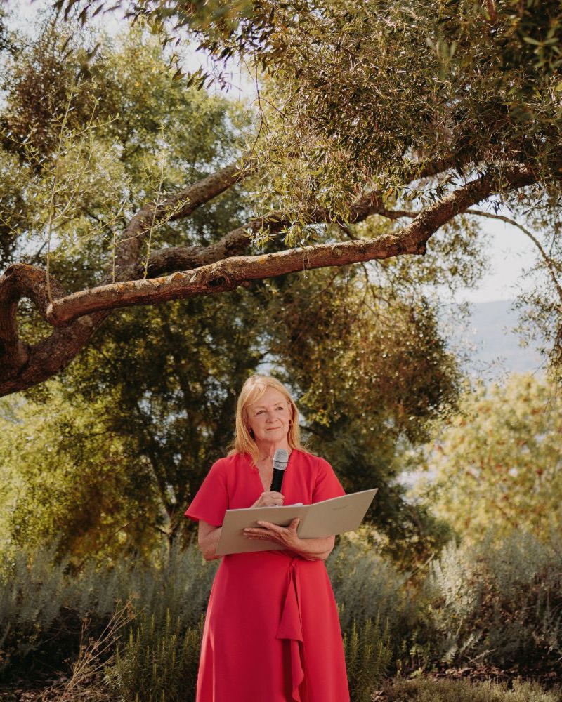 Chris in a red dress, leading an outdoor ceremony beneath an olive tree