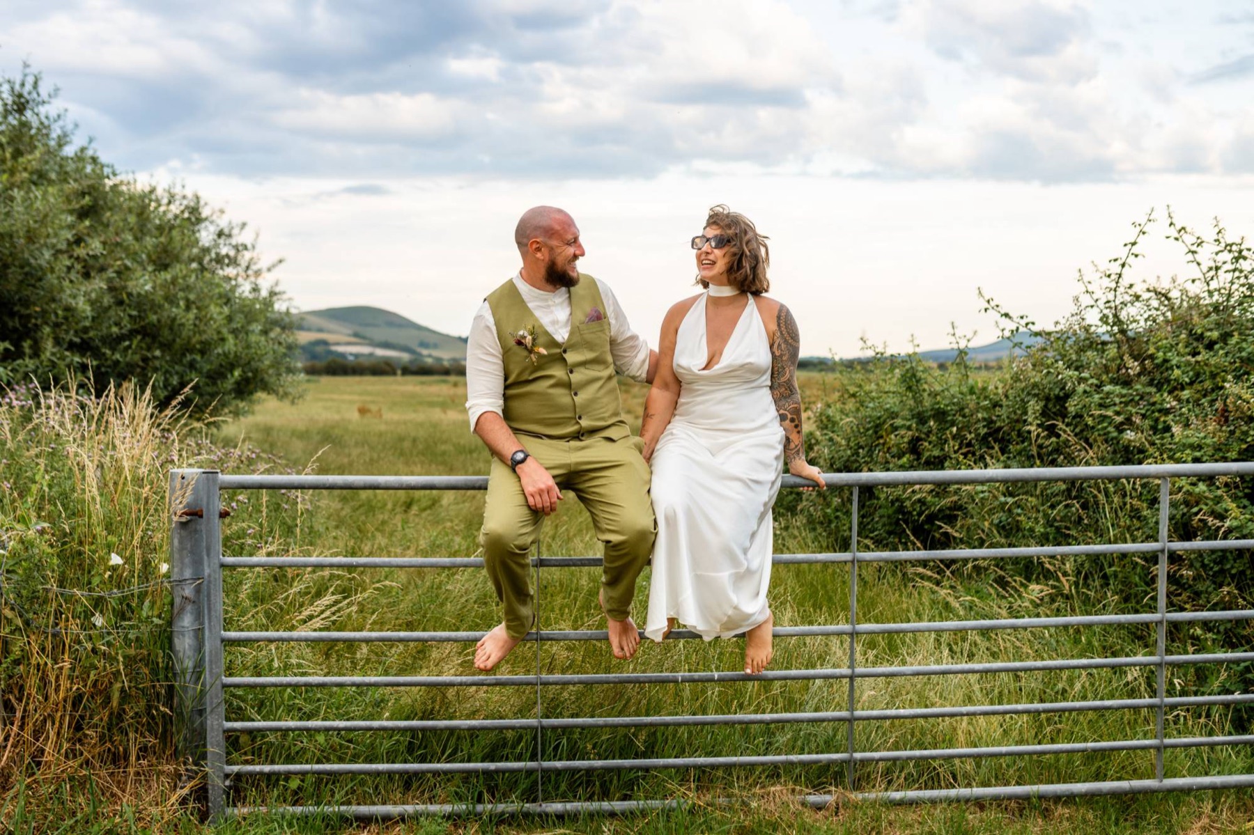 A newly married couple sitting barefoot on a field gate, laughing together, with hills behind them