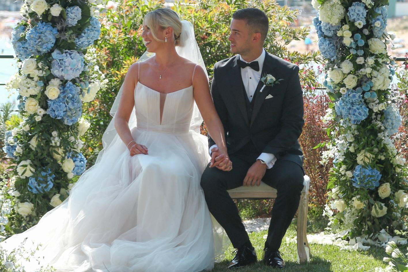 A bride and groom holding hands, seated beneath an arch of blue and white hydrangeas