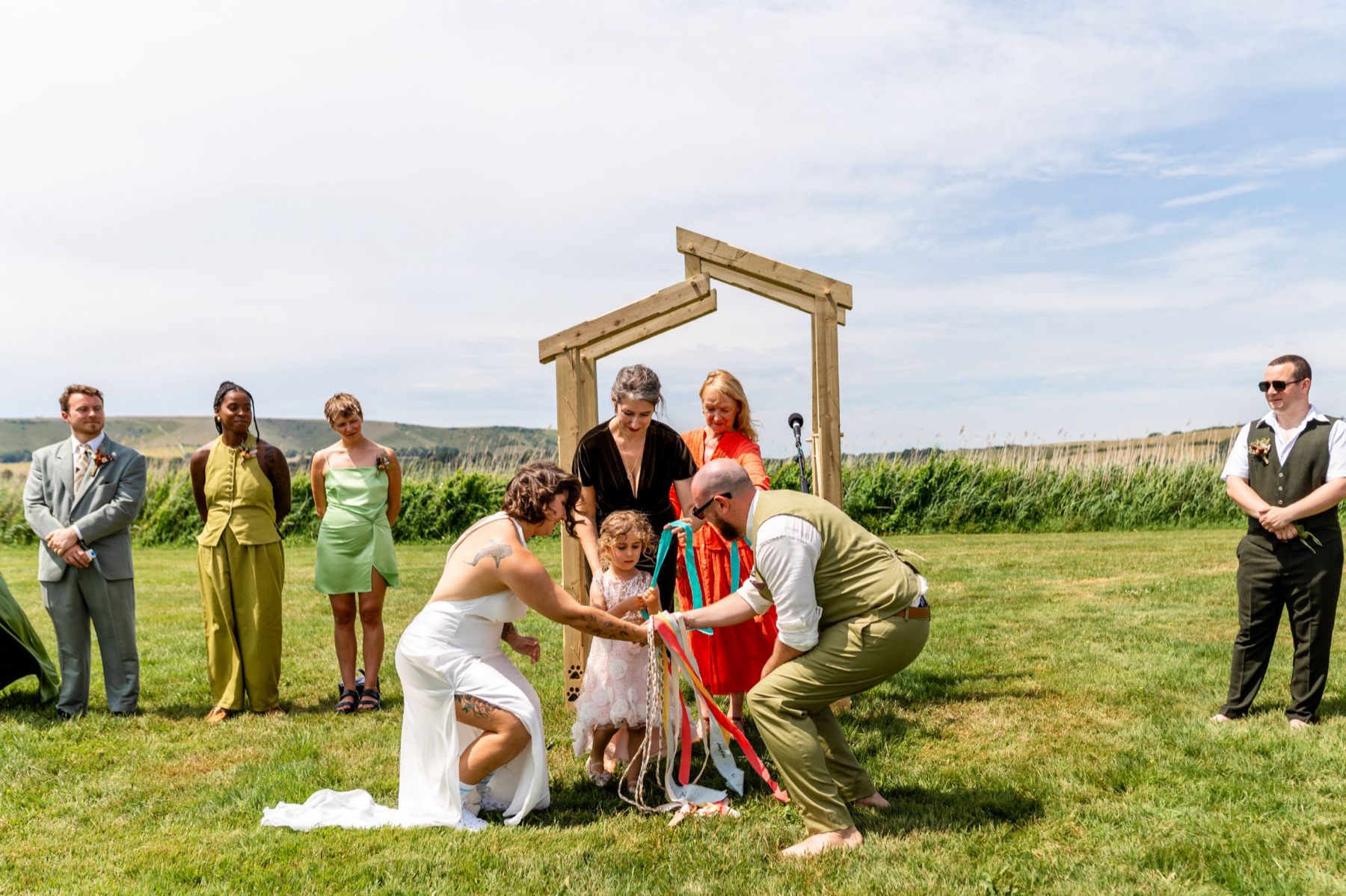 A couple's handfasting ceremony, with a young girl and family helping to tie colourful ribbons around their hands