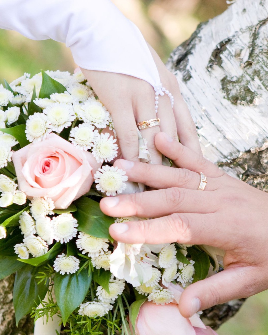 A couple's hands with wedding rings, resting on a bouquet of roses and white chrysanthemums